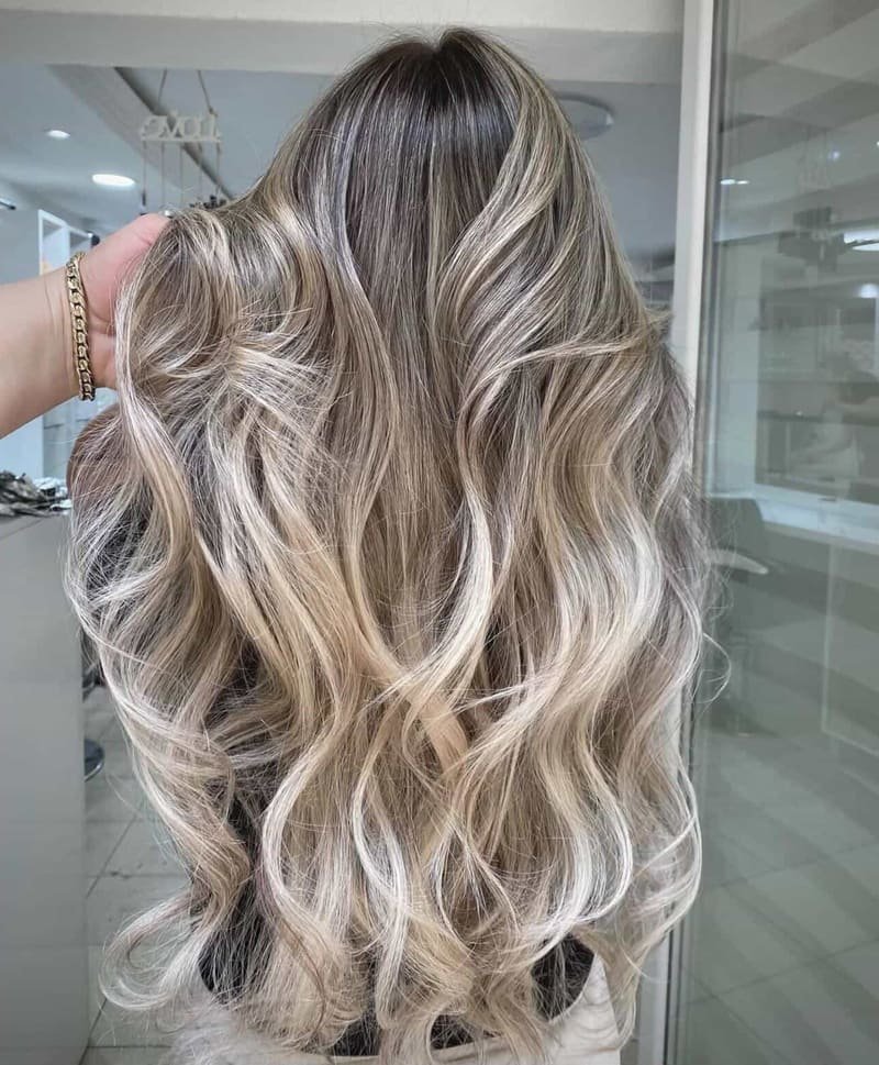 Ash blonde balayage with dark roots in long loose waves, photographed inside the studio