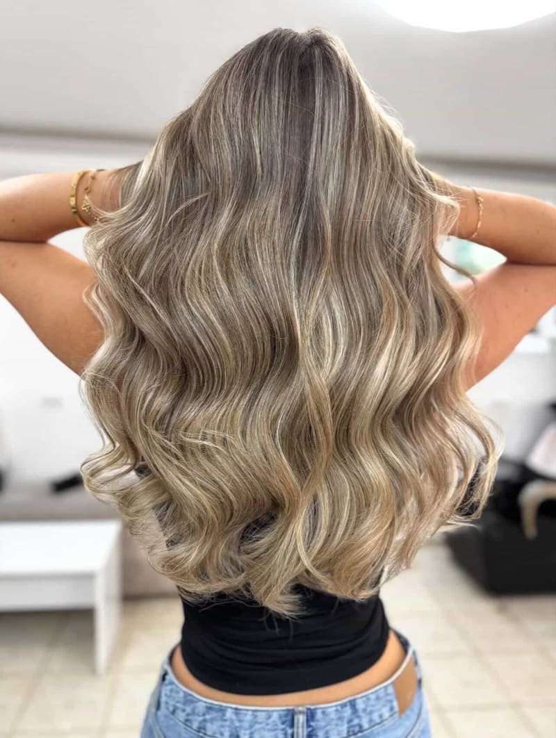 Beige blonde balayage in long glossy waves, with the arms raised behind the head