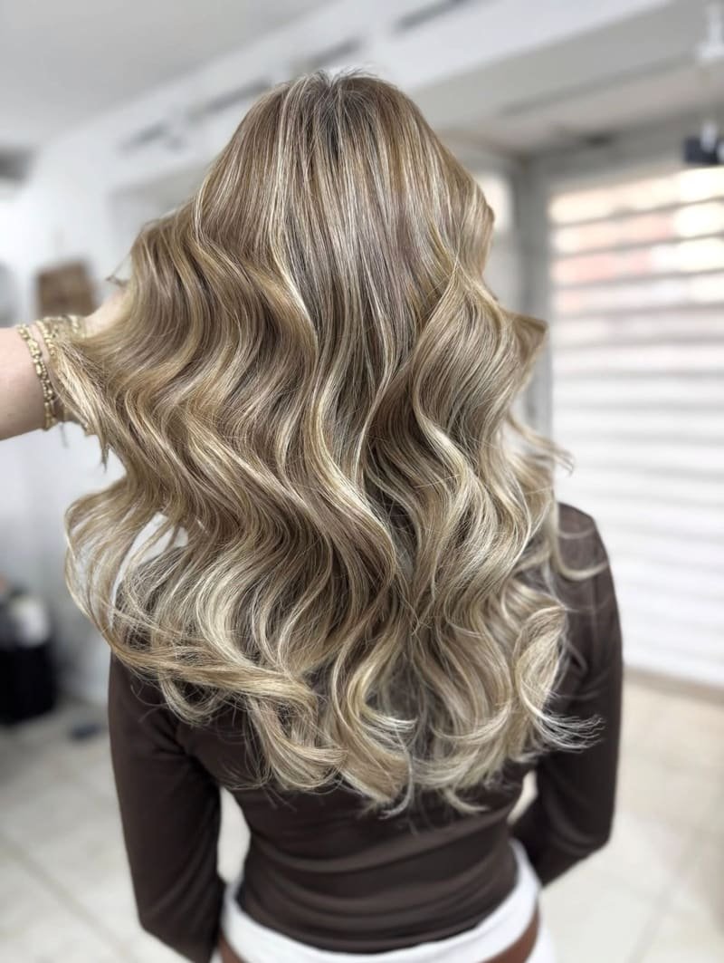Golden beige blonde highlights in long, defined waves, a hand with gold bracelets holding the hair back