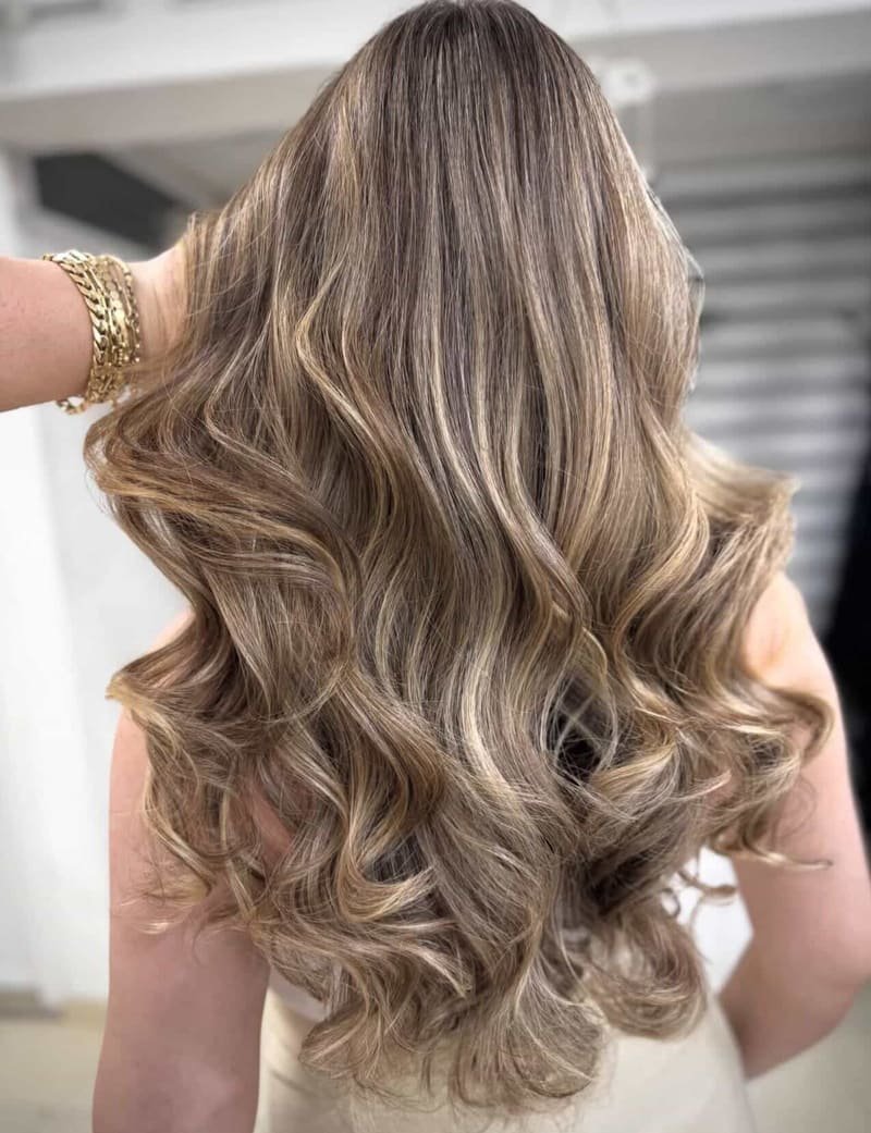 Long hair with honey and caramel balayage in soft waves, lifted by a hand wearing gold chain bracelets